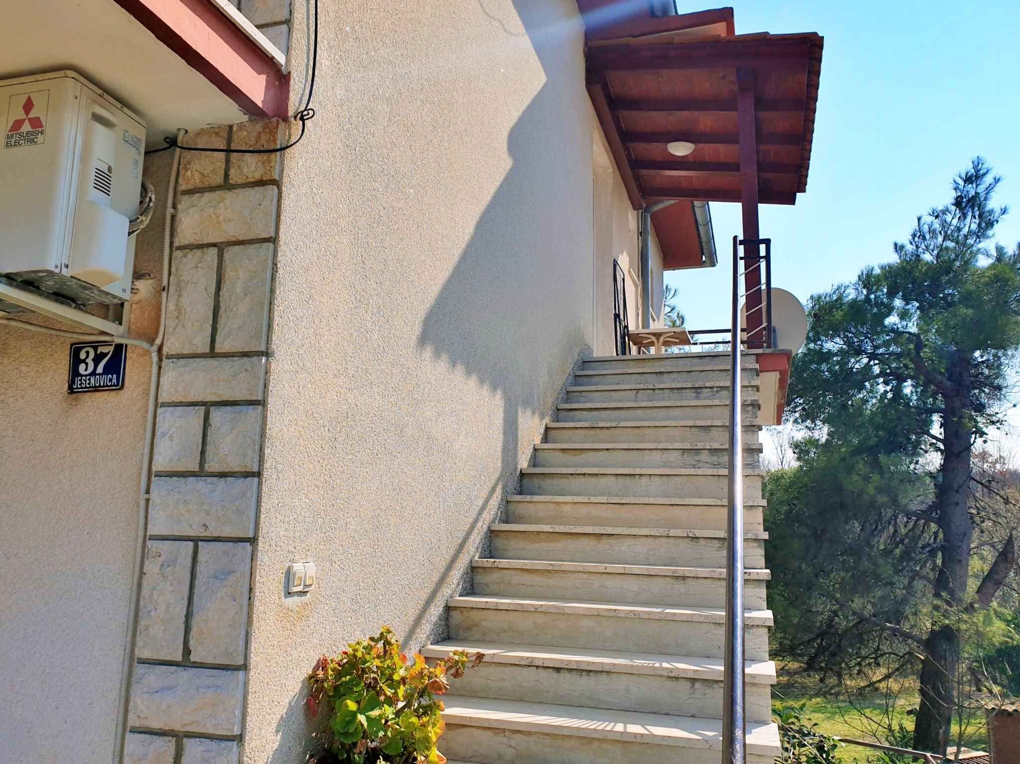 A residential building with a stone facade, a Mitsubishi electric air conditioning unit, and a wooden pergola on the upper floor.