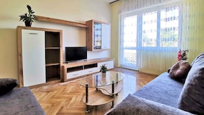Un salón con suelo de madera en espiga, una mesa de centro de cristal, un sofá gris, un televisor sobre un mueble de madera y una ventana con cortinas de encaje.