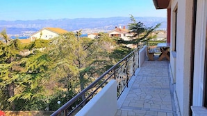 Un balcón con suelo de piedra, barandilla metálica y vistas a casas y árboles al fondo.