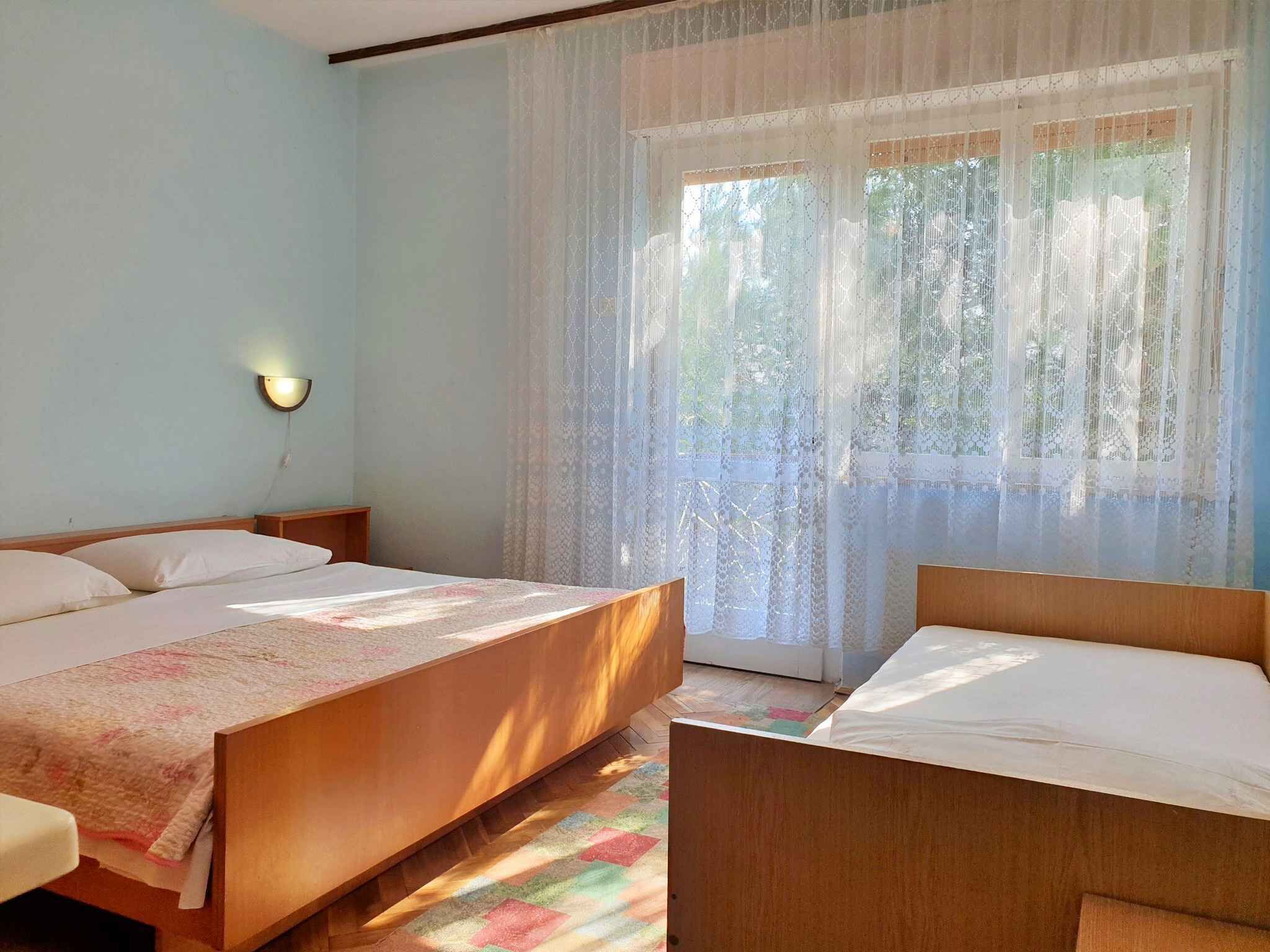 A room with two beds, a nightstand, a wall-mounted lamp, and a window with sheer curtains.