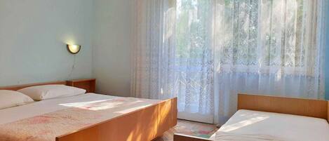 A room with two beds, a nightstand, a wall-mounted lamp, and a window with sheer curtains.