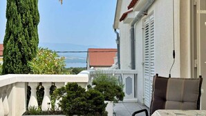 En balkon med udsigt over havet, et højt nåletræ og et lille bord med en stol.