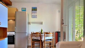 A compact kitchen with a refrigerator, stove, and dining table with chairs. A calendar is on the wall, and there is a window with blinds. - Holiday apartment with terrace and barbecue facilities (Šilo)