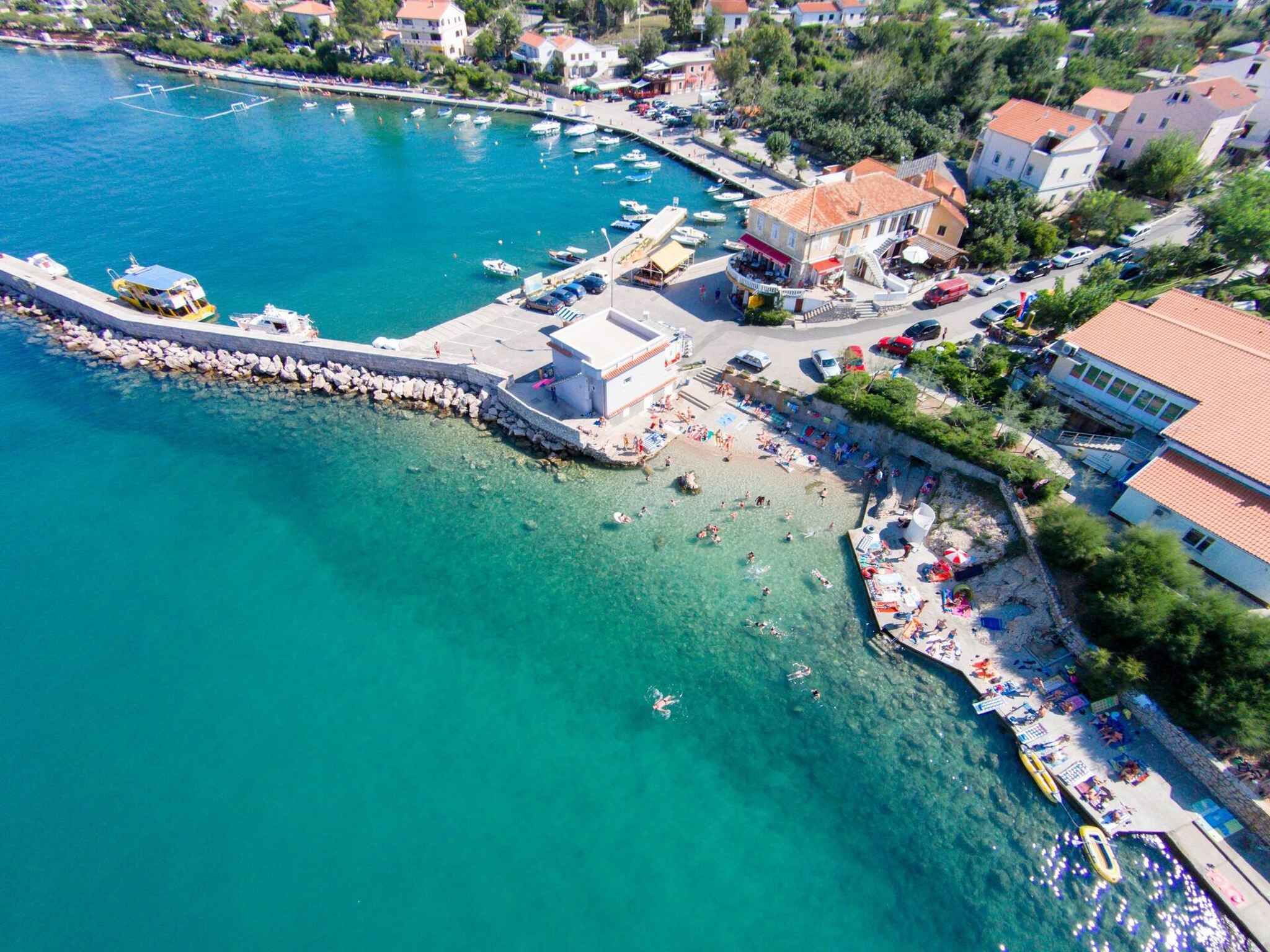 Luftaufnahme einer Küstenstadt mit einer Marina, klarem blauen Wasser und einem Strandabschnitt mit Menschen und Booten.