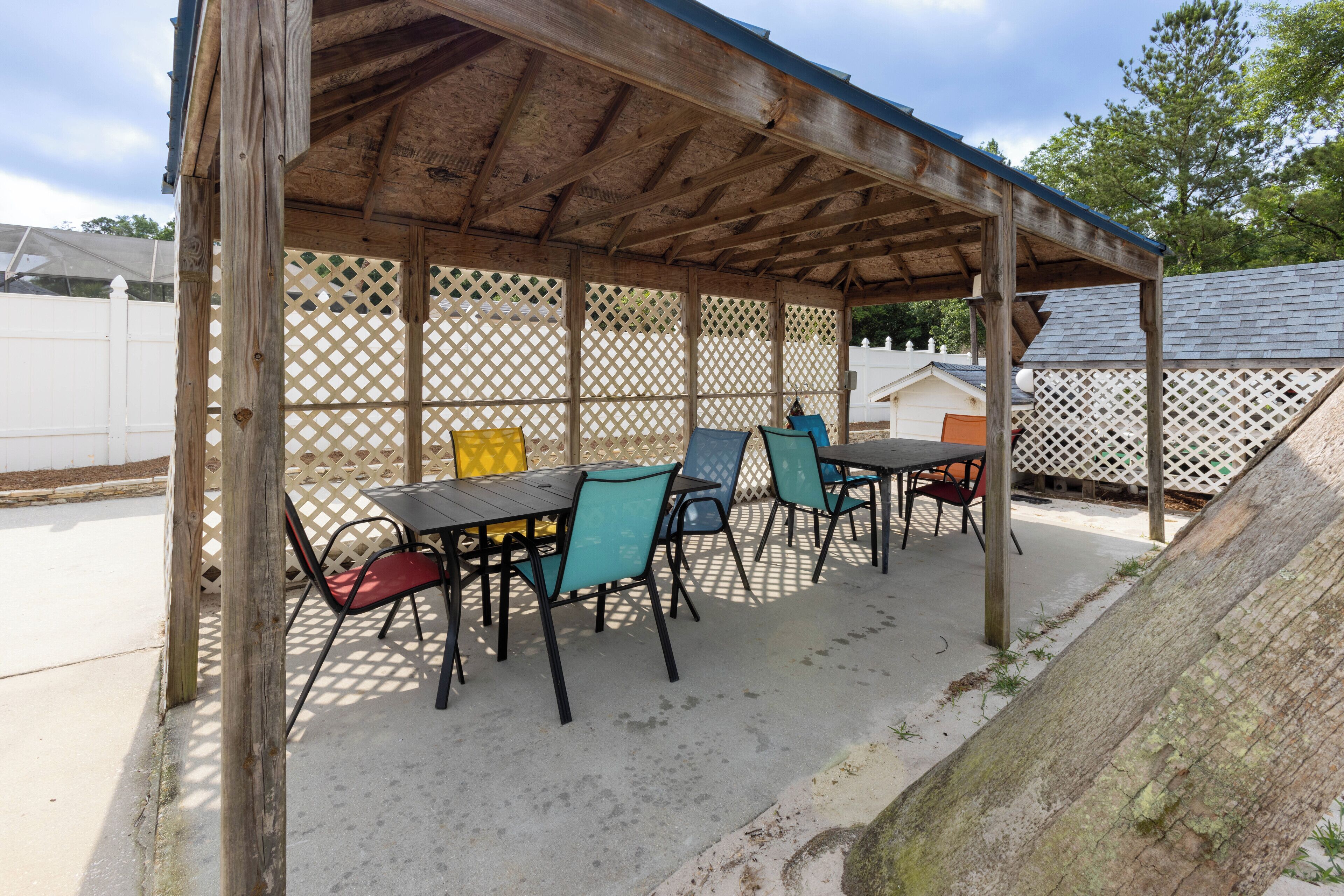 Een overdekt buitenzitgedeelte met tafels en stoelen, een houten pergola en een witte hek in de achtergrond.