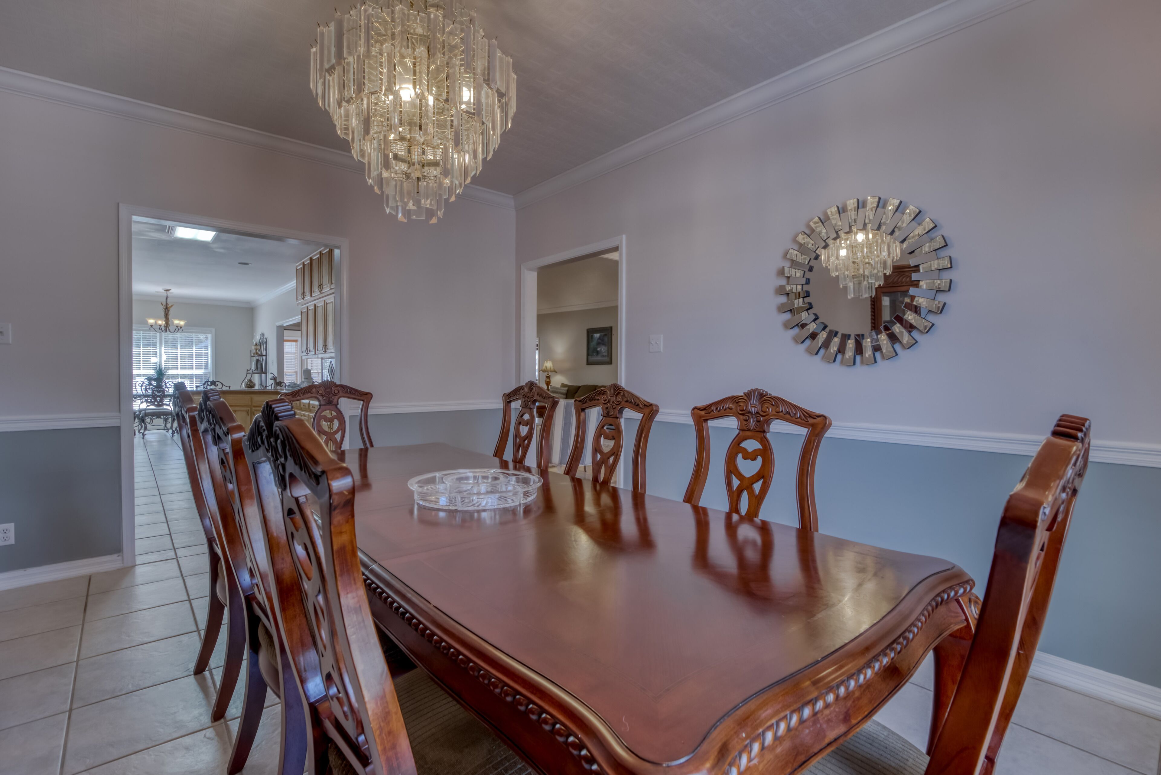 A dining room with a large wooden table, ornate chairs, a chandelier, and a mirror on the wall.