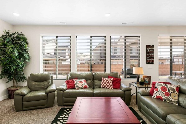 A living room with a sofa, armchair, and coffee table, large windows with blinds, and a potted plant. - Desert Rose, Newer Home, Community Pool! (Washington)