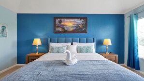 Ein Schlafzimmer mit einem großen Bett, zwei Nachttischen, einem schwanenförmigen Kopfkissen und einem Sonnenuntergangsbild an der Wand.