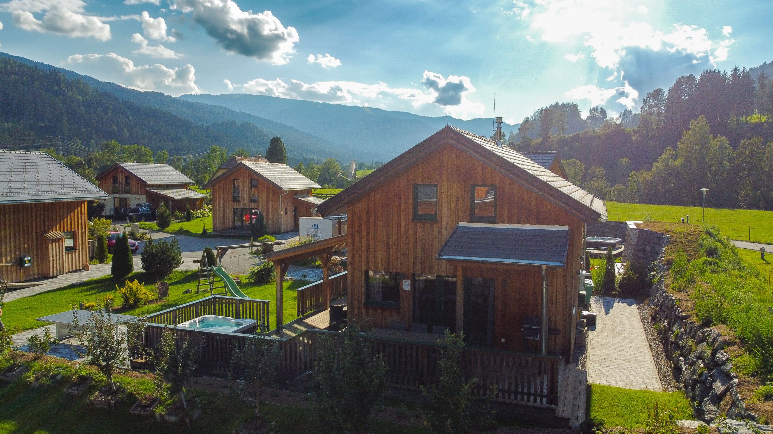 En træhytte med jacuzzi og terrasse, omgivet af frodigt grønt landskab og bjerge i baggrunden.