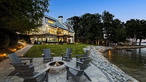 En patio ved søen med Adirondack-stole og et bålsted, med udsigt til et hus med en stor terrasse og en stenvæg.