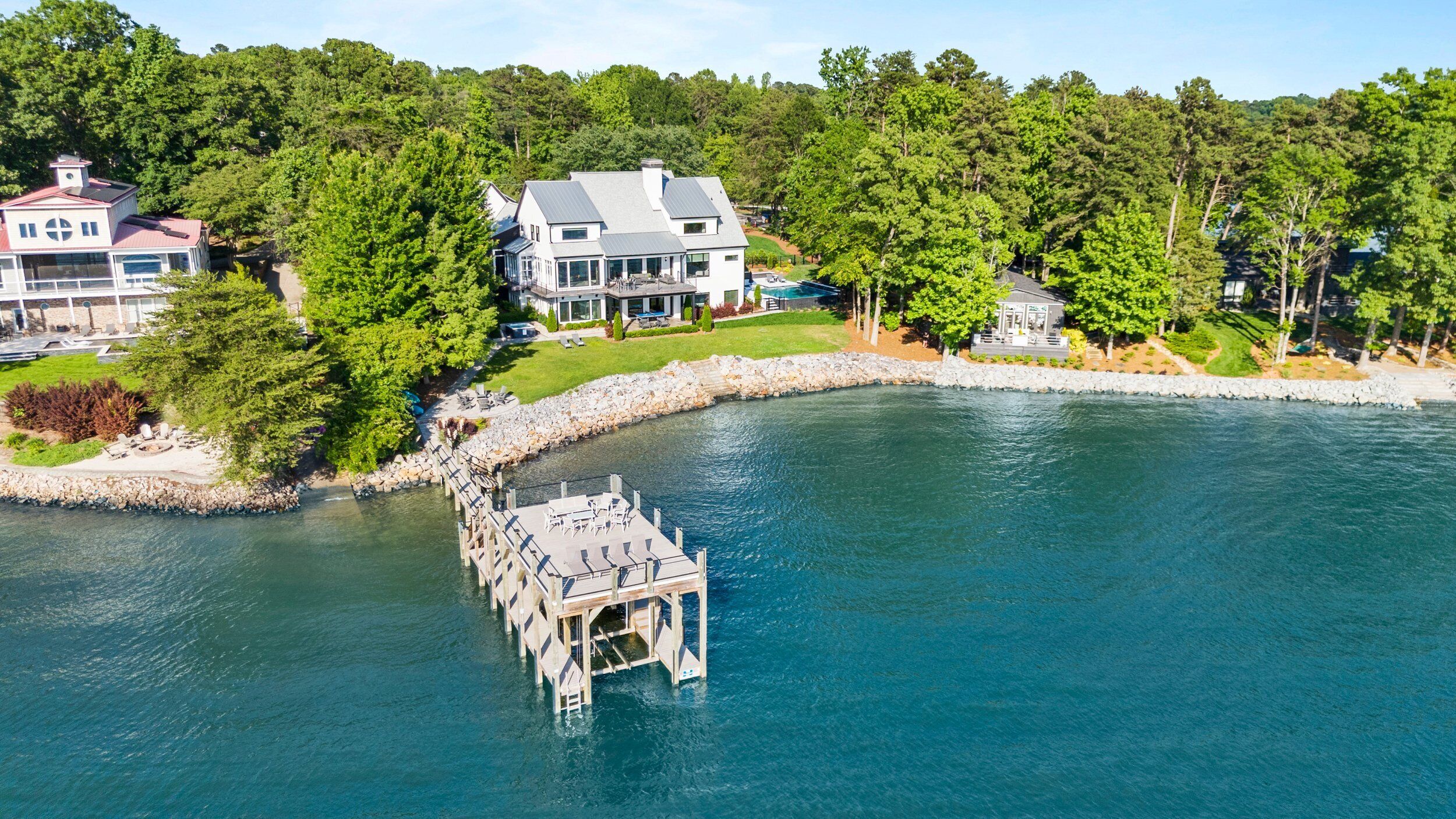 A waterfront property with a dock extending into a lake, surrounded by lush greenery and a large house with multiple windows and a covered patio.