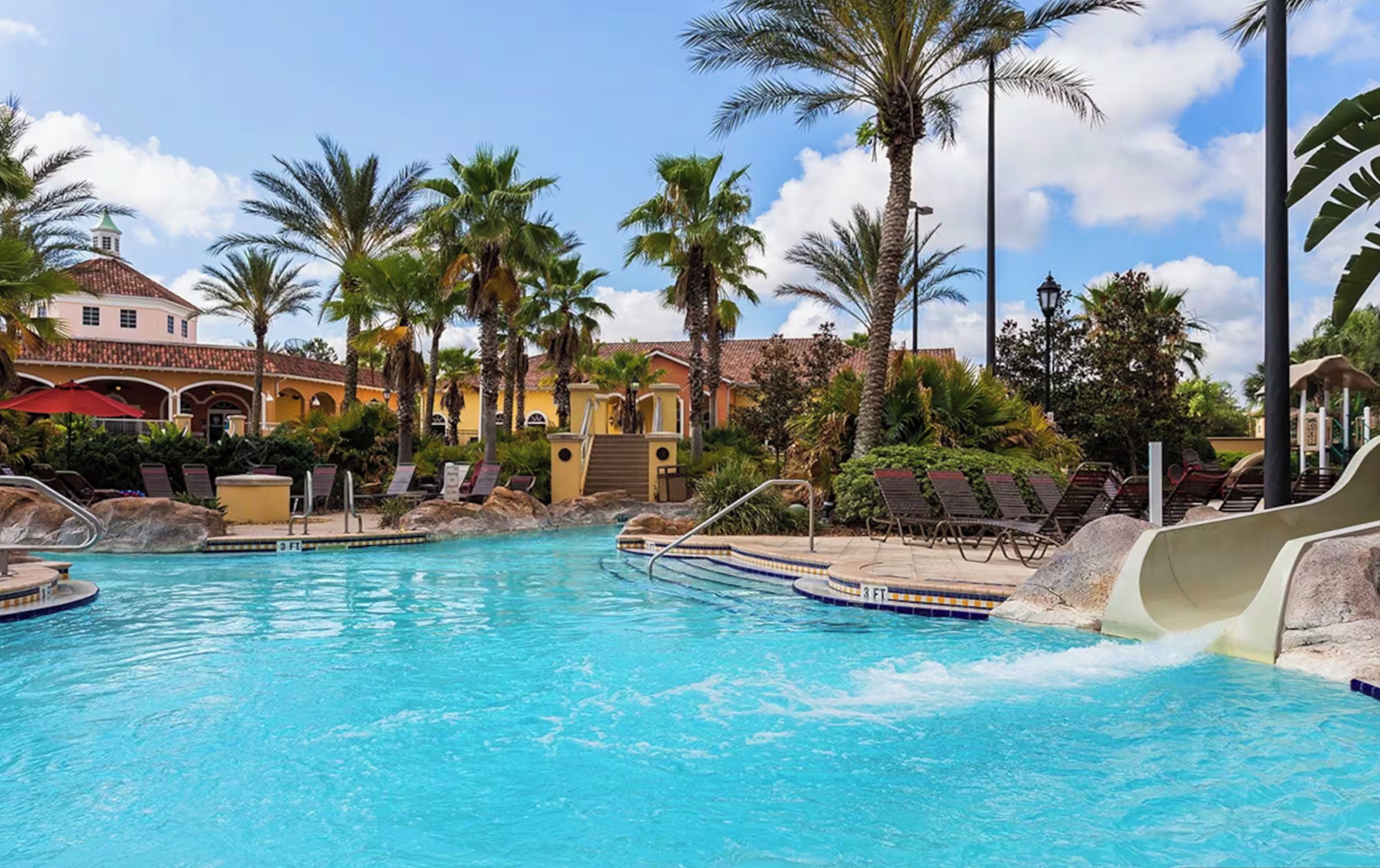 A swimming pool with a water slide, lounge chairs, palm trees, and a building with a tiled roof.