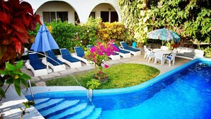 A swimming pool with lounge chairs and umbrellas, surrounded by greenery and a small garden. - Hotel Delfin (Barra de Navidad)