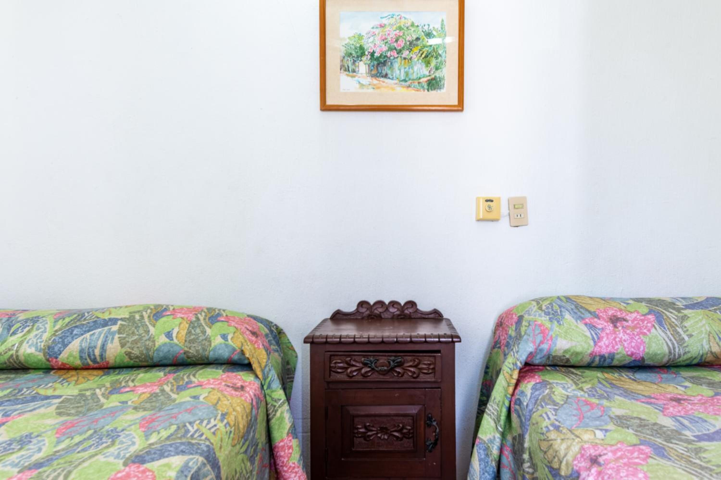 Habitació amb dos llits, cadascun amb una colcha de disseny floral, una petita taula de nit de fusta entre ells, i un quadre amb marc a la paret.