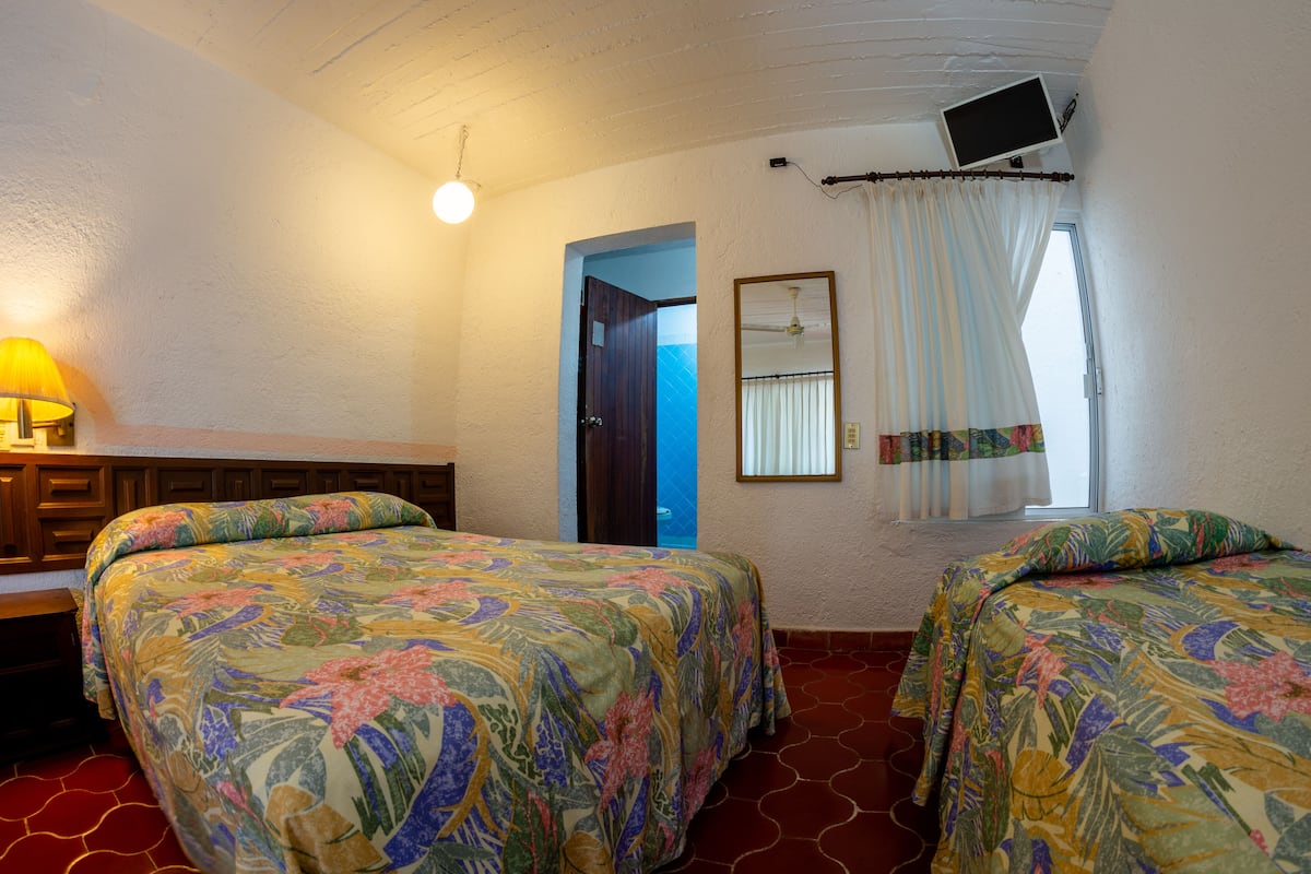 Una habitación con dos camas, un escritorio, una lámpara, un televisor montado en la pared y una ventana con cortinas.
