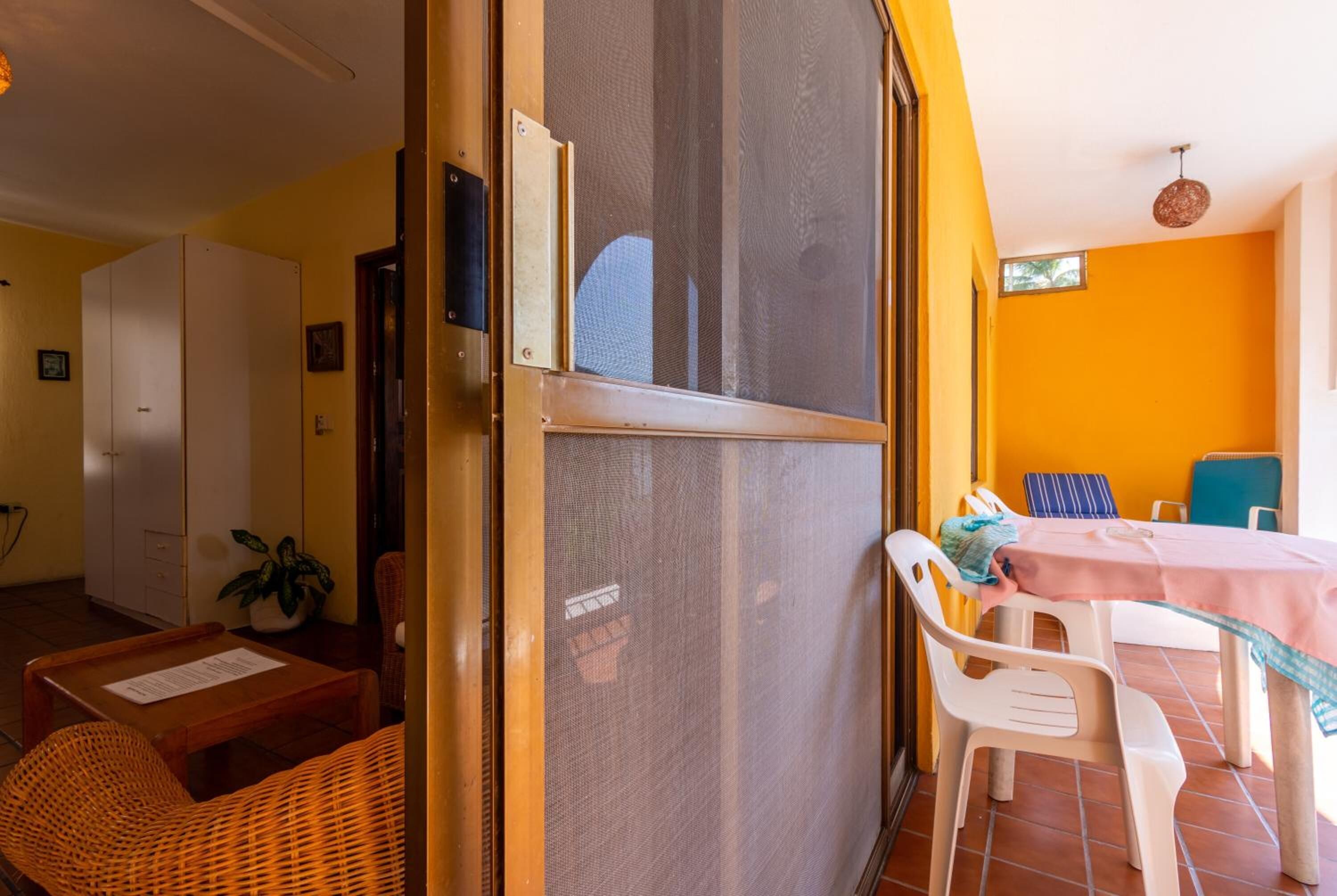 A room with a sliding glass door leading to a balcony, a small table with chairs, a wicker chair, and a white cabinet.