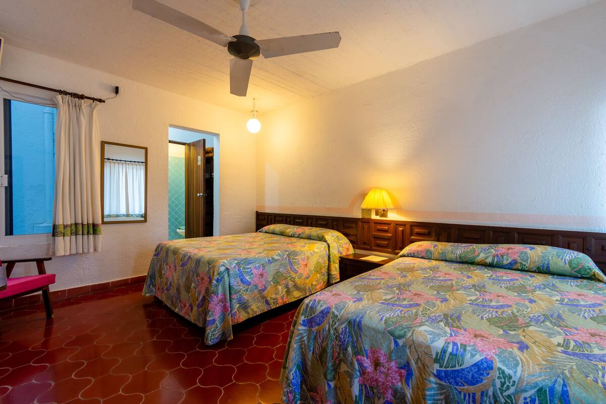 Habitación de hotel con dos camas, ventilador de techo, lámpara de noche y ventana con cortinas.