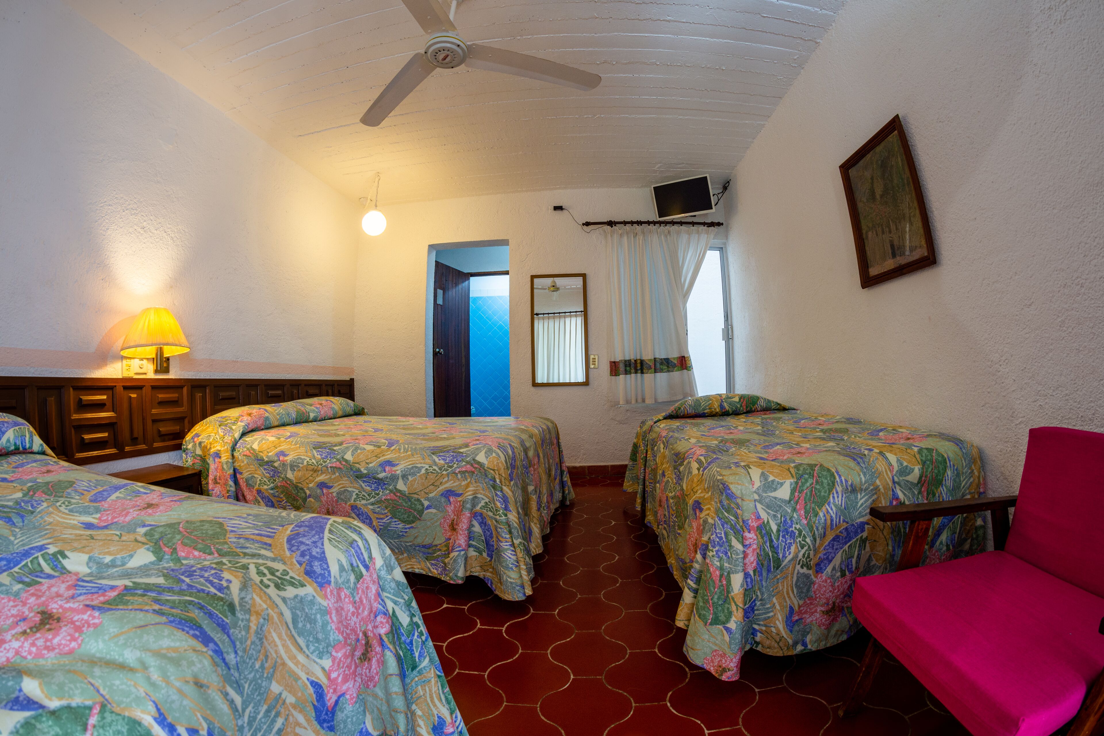 A room with two beds, a chair, a ceiling fan, a lamp, and a TV on the wall.