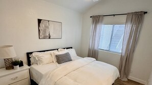 Une chambre à coucher comprenant un lit, une table de chevet, un ventilateur de plafond, une fenêtre avec des rideaux et un tableau abstrait accroché au mur.