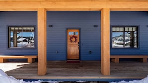 A wooden porch with a blue exterior, a wooden door with a wreath, and two windows on either side. - Big Sky Escape - Spacious Home with Hot Tub (Big Sky)