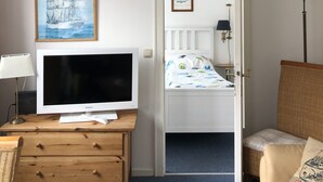 A room with a wooden desk, a television, a bed with a white headboard, a wicker chair, and a lamp. - Bright, cozy vacation apartment in a perfect location close to the beach and town center  (Sylt)
