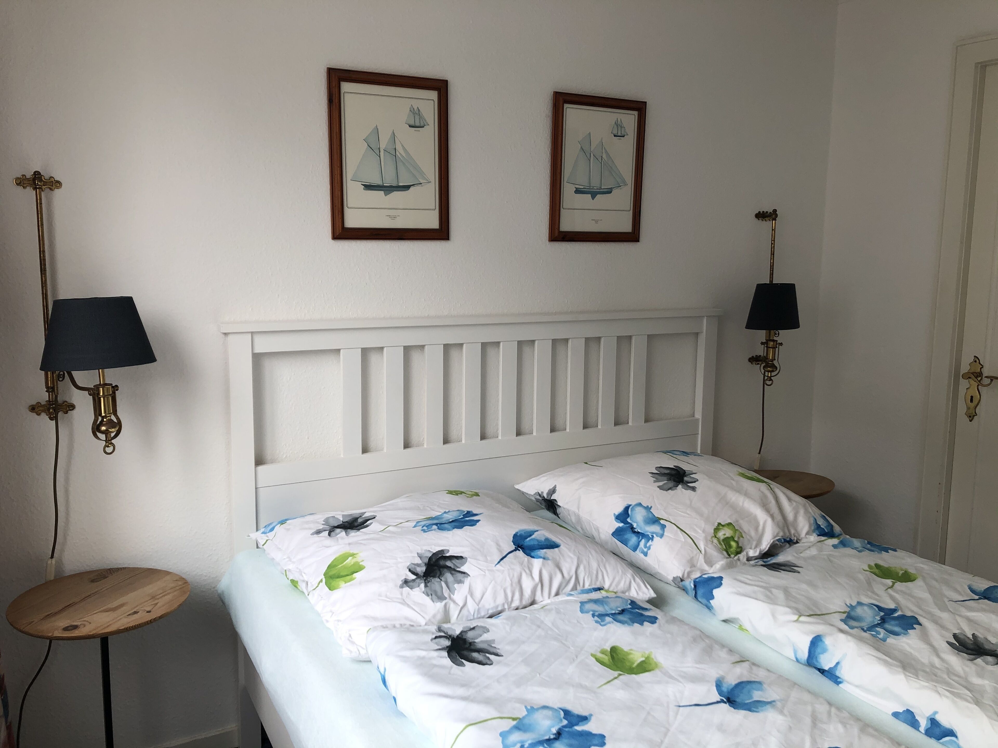 A bedroom with a white headboard, floral bedding, two framed pictures on the wall, and two bedside lamps.