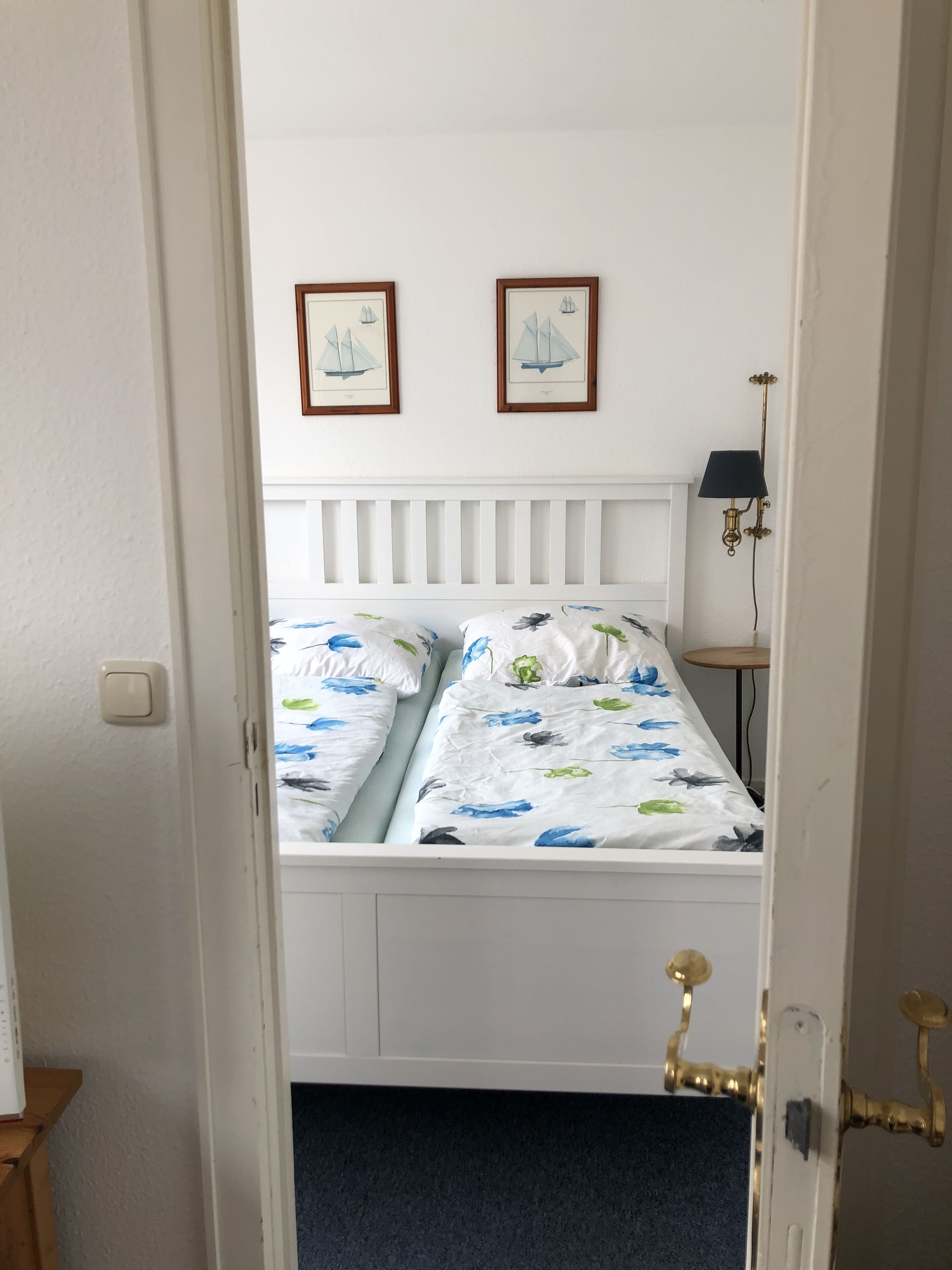 Un dormitori amb un llit blanc, roba de llit amb motius florals, dos quadres a la paret, una làmpada de nit i una taula de fusta petita.