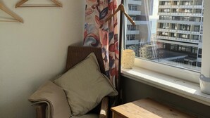 A corner room with a wicker chair, a wooden dresser, a window with a view of a building, and a floor lamp. - Bright, cozy vacation apartment in a perfect location close to the beach and town center  (Sylt)