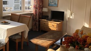 A hotel room with a dining table, chairs, a television, a wooden cabinet, a desk, a lamp, a window with curtains, a painting, and a vase with flowers. - Bright, cozy vacation apartment in a perfect location close to the beach and town center  (Sylt)