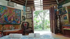 Une pièce avec un plafond haut, un grand tableau aux couleurs vives, une chaise, une table et une vue sur la verdure à l’extérieur.