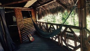 Une véranda en bois, avec un hamac et une guitare, entourée de structures en bambou et surmontée d’un toit de chaume.