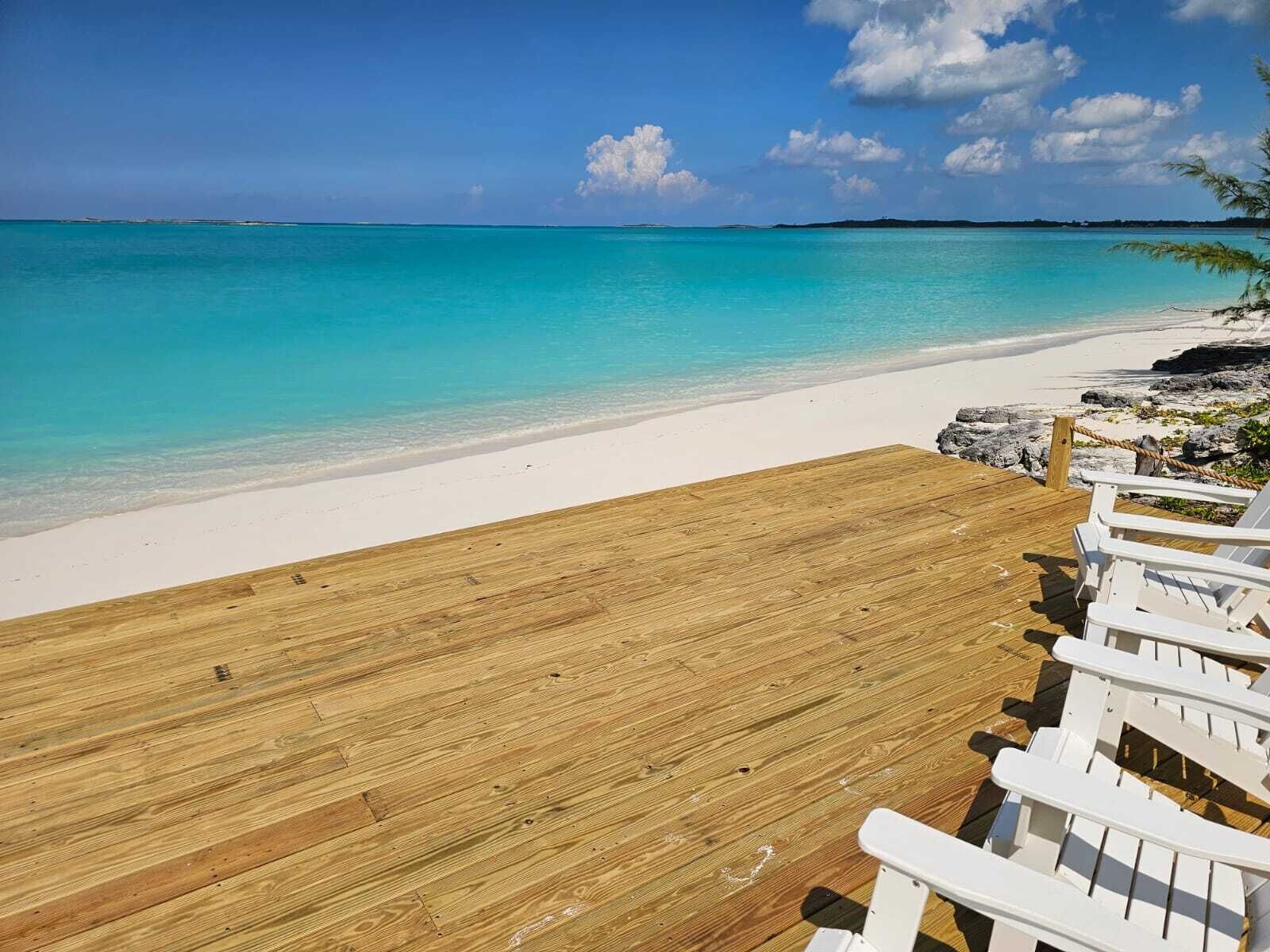 Eine Holzterrasse mit weißen Stühlen mit Blick auf einen Strand mit klarem türkisblauem Wasser und weißem Sand.