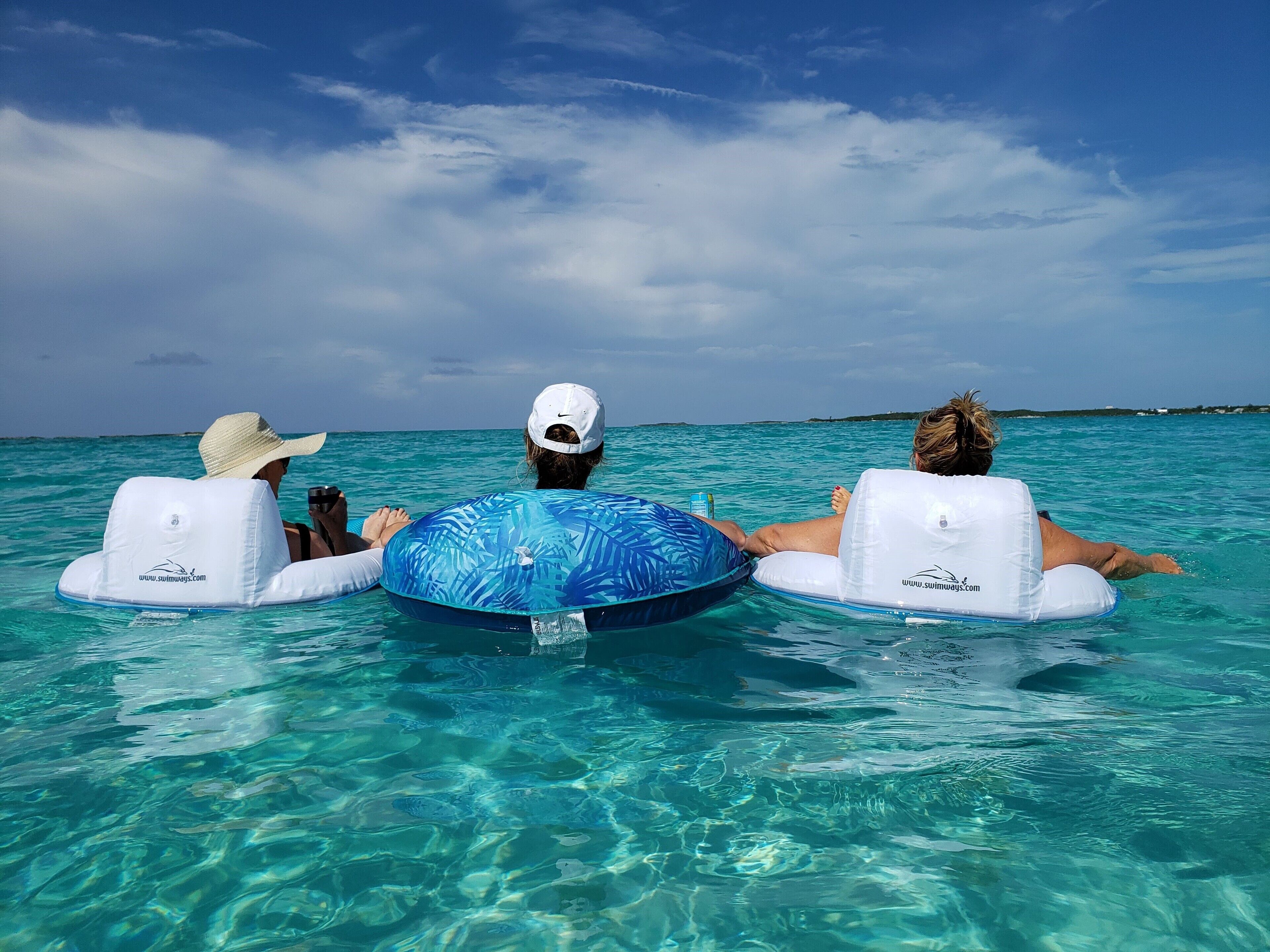 Tres personas flotando en el agua con flotadores circulares, agua turquesa cristalina y una orilla lejana.