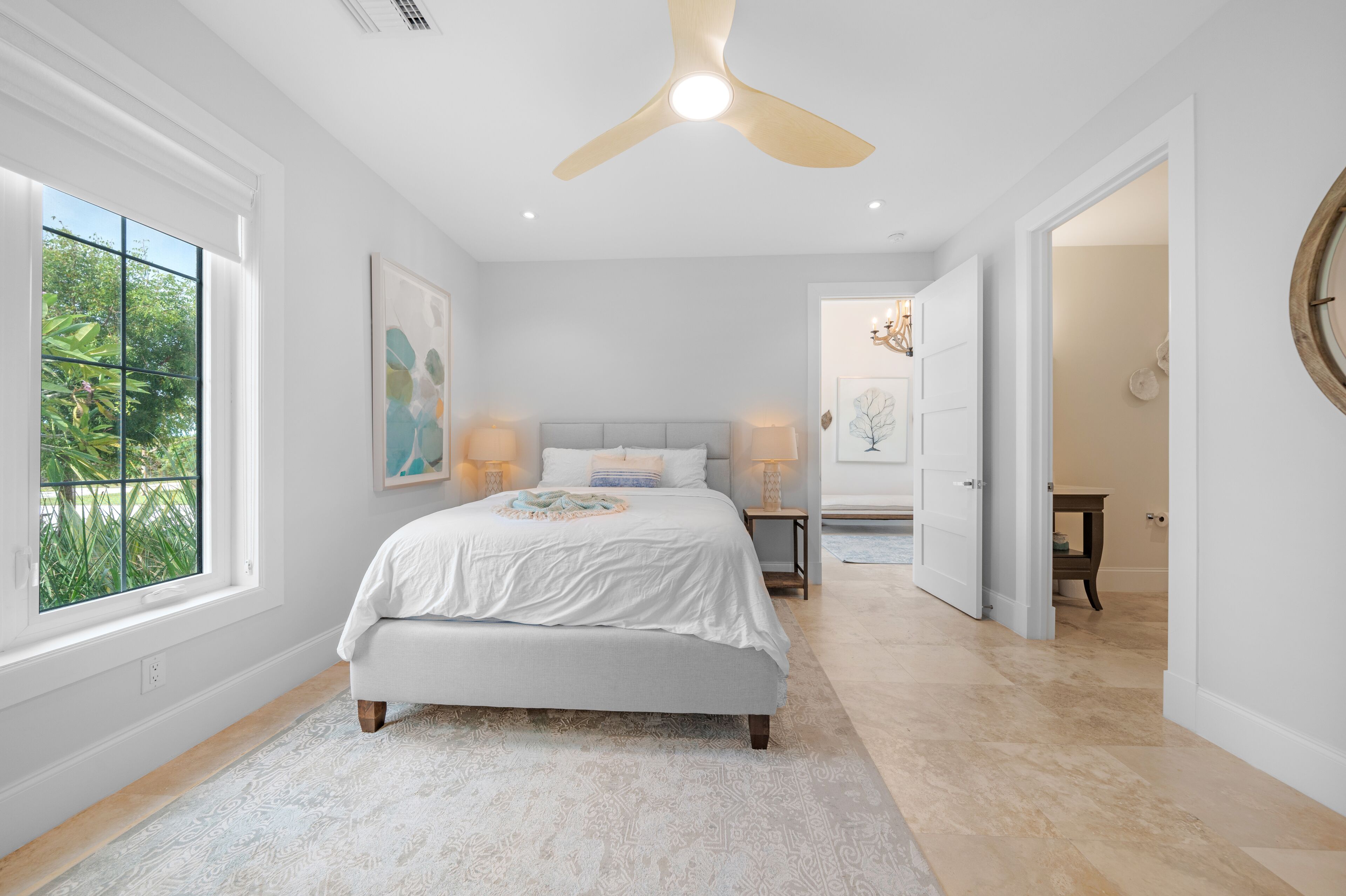 A bedroom with a bed, bedside lamps, a ceiling fan, and a window with a view of greenery.