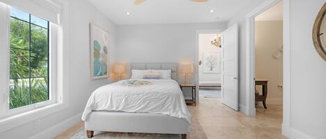 A bedroom with a bed, bedside lamps, a ceiling fan, and a window with a view of greenery.