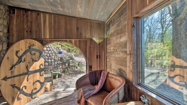 Ein rustikales Blockhaus-Interieur mit einer Steinwand, einer Holztür mit Metallverzierungen, einem Ledersessel und einem Fenster mit Blick auf Bäume.