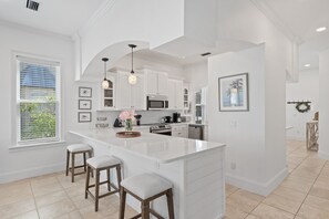 Una cocina moderna con armarios blancos, una isla grande con taburetes de bar, iluminación colgante y vistas al exterior a través de una ventana con persianas.