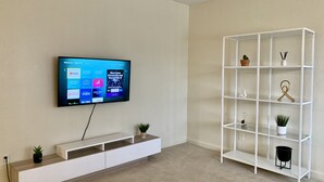 Una sala de estar moderna con un televisor de pantalla plana montado que muestra servicios de streaming, un mueble blanco para entretenimiento con una planta y una estantería blanca con objetos decorativos.