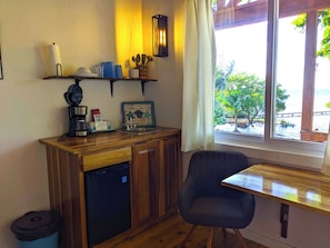 A compact kitchen with a dishwasher, a coffee maker, and a sink. There's a chair and a table near a window with a view of greenery and a dock.