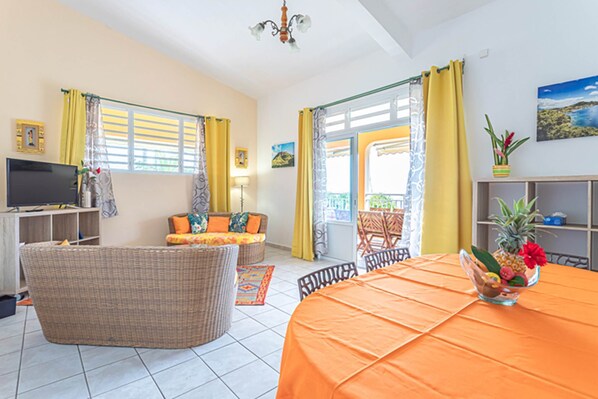 A living room with a television, wicker furniture, and a dining table with an orange tablecloth. - FLAMBOYANT (3 - 8 people under 12 years old) (DESHAIES)