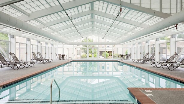 Una amplia piscina interior con paredes y techo de vidrio transparente, rodeada de sillones y mesas.
