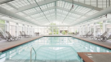 Une grande piscine intérieure aux parois et au plafond en verre transparent, entourée de chaises longues et de tables.
