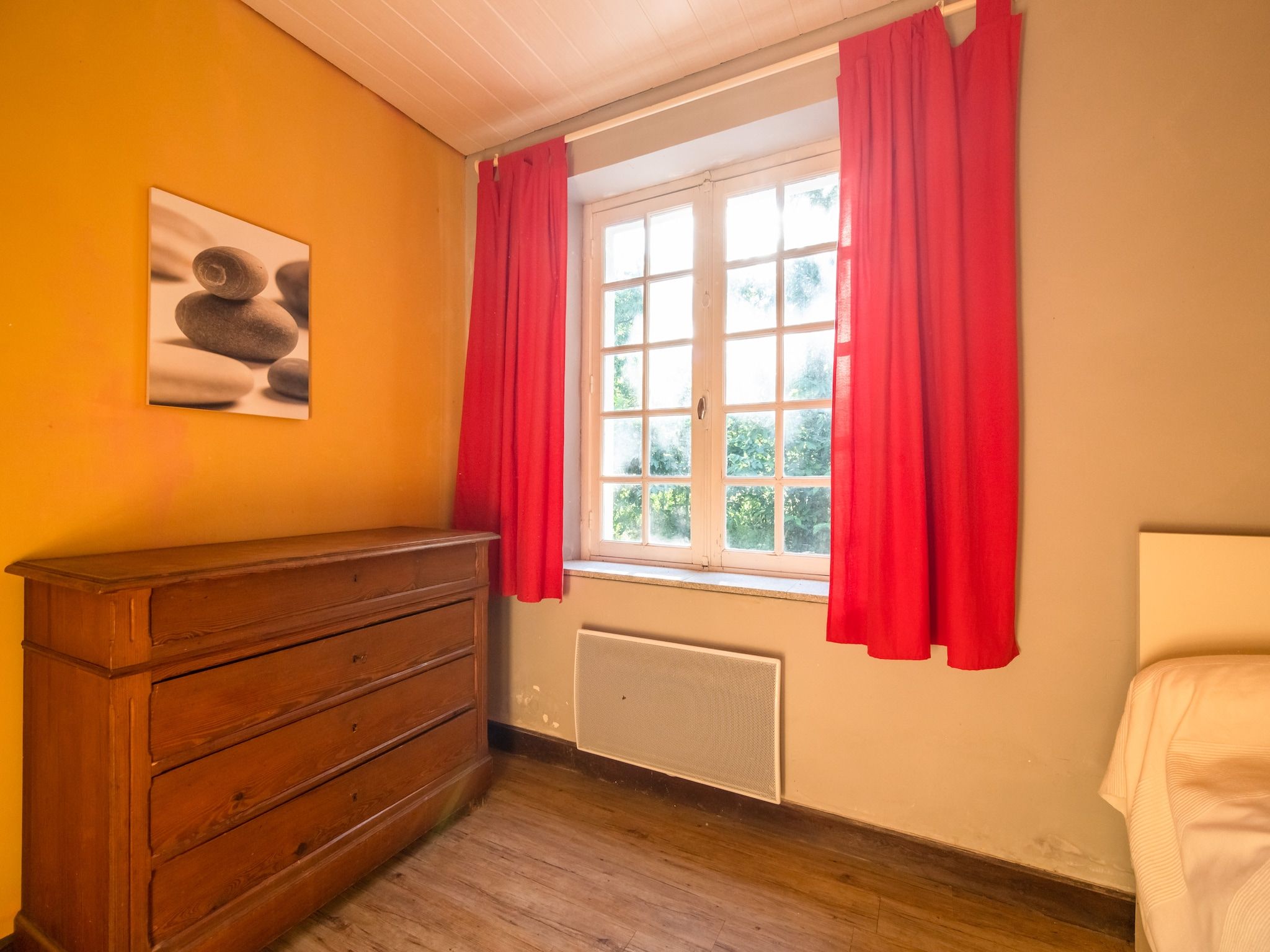 Una habitación con un aparador de madera, un radiador, una ventana con cortinas rojas y un cuadro en la pared.