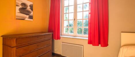 Una habitación con un aparador de madera, un radiador, una ventana con cortinas rojas y un cuadro en la pared.