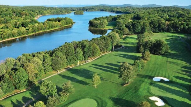 Vista aérea de un campo de golf con un lago, rodeado de densos bosques y onduladas colinas.