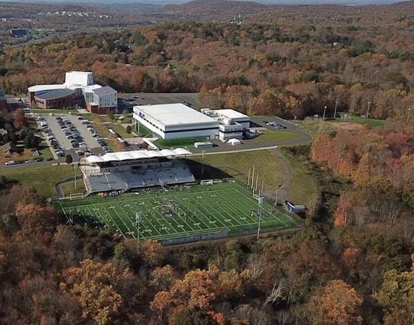 An aerial view of a campus with a football field, buildings, and surrounding wooded area.