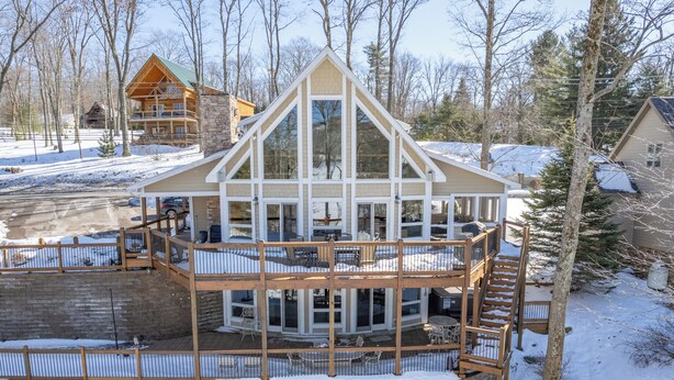 A multi-story lodge with a central glass block structure, wooden deck, and a stone retaining wall. - Stylish Lakefront Home w/ Foosball & Hot Tub (Oakland)