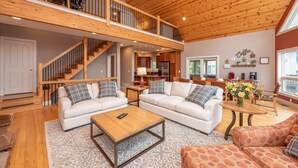 A spacious living area with two white sofas, a wooden coffee table, and a patterned rug. A staircase leads to an upper level with a balcony. - Stylish Lakefront Home w/ Foosball & Hot Tub (Oakland)