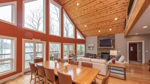 A spacious living area with a wooden ceiling, large windows, a fireplace, and a television. - Stylish Lakefront Home w/ Foosball & Hot Tub (Oakland)