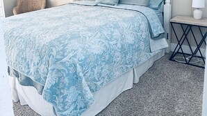 Un dormitorio con cama blanca, ropa de cama azul claro, un espejo redondo y una mesita de noche pequeña.